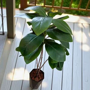 以下が含まれることがあります： 大きな光沢のある緑の葉を持つ、鉢植えの人工植物。植物には複数の茶色の茎があり、茶色のマルチで満たされた黒い幾何学的な形の鉢に植えられています。植物の高さは約60cmです。