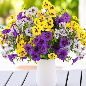 Puede incluir: Un vibrante ramo de pensamientos artificiales en un jarrón blanco. Las flores son una mezcla de amarillo, morado y blanco, con follaje verde. El jarrón está sobre una mesa de madera blanca, con un fondo verde borroso.