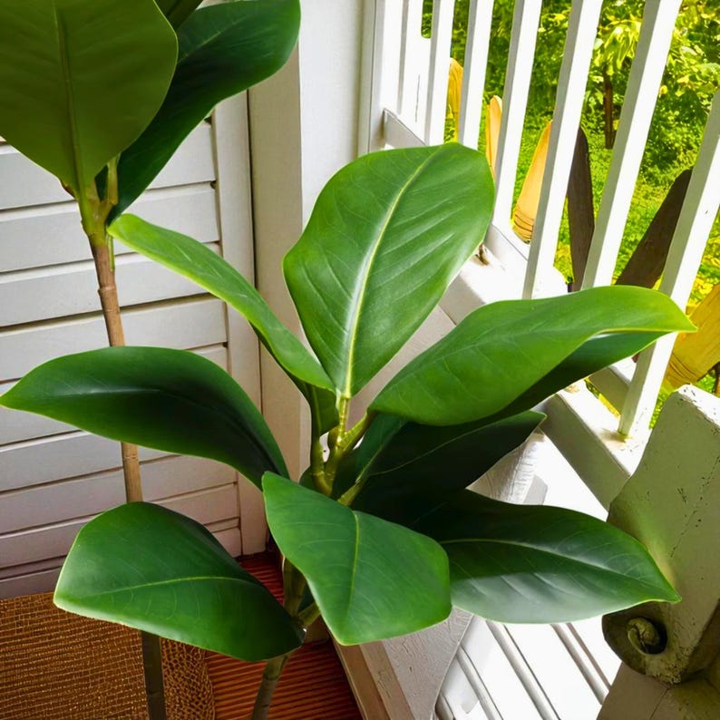 以下が含まれることがあります： 鮮やかな緑色の人工植物。大きく光沢のある葉があり、茶色の茎が特徴です。白い手すりの近くに配置されています。葉には目立つ中心静脈があり、植物のリアルさを高めています。