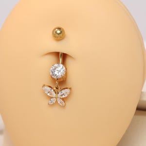 May include: Gold-coloured belly button ring with a dangling butterfly charm. The charm is decorated with clear, sparkling crystals. The top and bottom balls are gold. The jewellery is displayed on a tan-coloured surface.