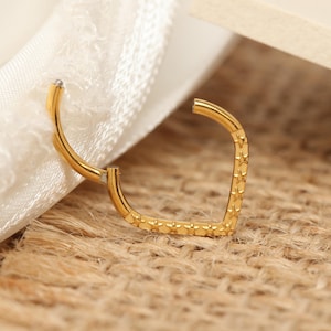 Puede incluir: Un pendiente dorado en forma de V con una superficie texturizada. El pendiente está abierto, revelando el mecanismo de cierre. El fondo incluye una tela blanca y una superficie texturizada de color beige.