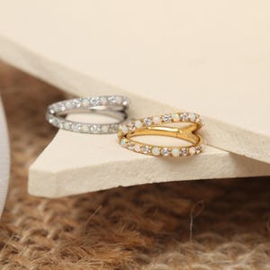 May include: Two rings, one silver and one gold, each with a double band design. Both rings are adorned with small, round, white gemstones. The gold ring has a warm, lustrous finish, while the silver ring has a bright, polished appearance.