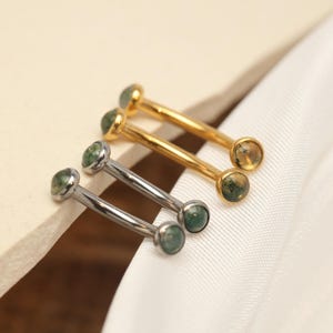 May include: Close-up of four body jewelry pieces. Two are silver-toned, and two are gold-toned. Each piece has a straight bar with a small, round, green stone at each end. The jewelry is displayed on a white, textured surface.
