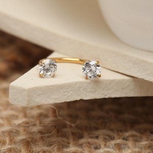 May include: A gold-colored, open-hoop earring with two clear, round, sparkling gemstones. The earring is displayed on a white, angled surface against a neutral background. The design is simple and elegant.