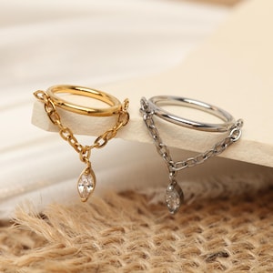 Puede incluir: Dos anillos, uno dorado y otro plateado, cada uno con una cadena y una gema transparente en forma de lágrima colgante. Los anillos se exhiben sobre una superficie neutra con un fondo texturizado y natural.