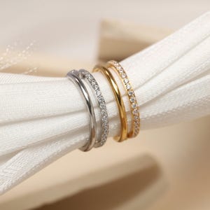 Puede incluir: Tres anillos, dos plateados y uno dorado, se muestran sobre una tela blanca. Los anillos plateados tienen un diseño de doble banda, con una banda incrustada con pequeñas piedras transparentes. El anillo dorado también tiene un diseño de doble banda, con una banda incrustada con pequeñas piedras transparentes.