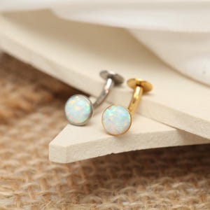 May include: Two opal stud earrings. One earring has a silver post and setting, the other has a gold post and setting. The opal stones are round and have a white and iridescent appearance. The earrings are displayed on a neutral surface.