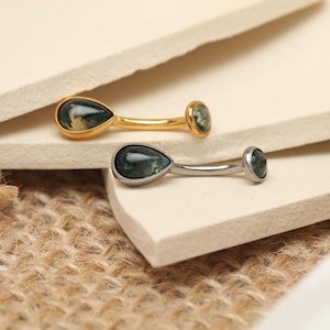 Pode incluir: Duas peças de joalharia, uma dourada e outra prateada, cada uma com uma pedra verde em forma de gota. A peça dourada tem uma pedra redonda a condizer na extremidade da barra. As joias repousam numa superfície de tom neutro com textura de serapilheira.