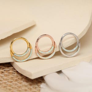 May include: Three metal hoop earrings in gold, rose gold, and silver. Each earring features a double-layered design with a curved, opal-like inlay. The earrings are displayed on a neutral-toned surface, showcasing their different finishes.
