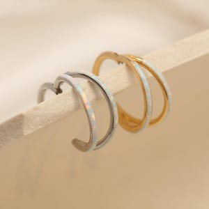 Op de afbeelding: Twee paar oorringen. Eén paar is zilverkleurig en het andere is goudkleurig. Beide paren hebben een dubbel ringontwerp met een witte, opaalachtige inleg. De oorbellen worden gepresenteerd op een lichtgekleurd oppervlak.