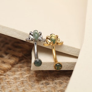 May include: Two curved eyebrow rings. One is silver-toned, the other gold-toned. Each features a frog-shaped design with a green stone at the top and a matching green stone at the bottom. The frog's body is textured, and the stones have a mottled appearance.