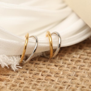 May include: Two double hoop earrings, one gold-coloured and one silver-coloured, are presented on white fabric. The earrings feature a minimalist design. The background is a textured, neutral-toned surface.