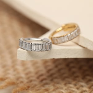 May include: Two rings, one silver and one gold, each adorned with a row of rectangular, clear gemstones. The rings are set against a neutral background, highlighting the sparkle of the stones. The gold ring is in the background, while the silver ring is in the foreground.