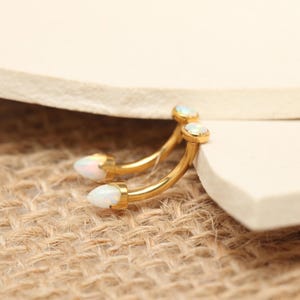 May include: Two gold-colored, curved eyebrow rings with white opal-like ends. The jewelry is set against a neutral background, highlighting the curved design and the iridescent quality of the opal stones. The curved shape is ideal for eyebrow piercings.