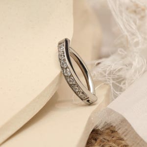 May include: A silver belly button ring, curved in shape, adorned with a line of small, clear gemstones. The ring's polished metal and sparkling stones are set against a neutral backdrop. A delicate piece of body jewellery.