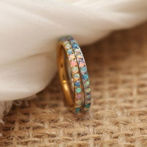 May include: Two gold rings with a band of small, colorful opals. The opals display a range of colors, including pink, blue, and white. The rings are set against a neutral background, highlighting the jewelry's design.