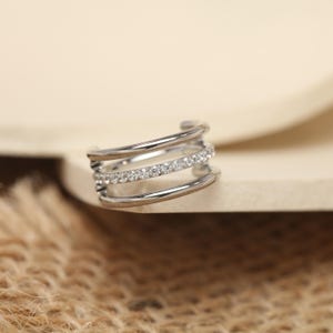 May include: A silver-toned ear cuff featuring three stacked bands. The middle band is adorned with a row of small, clear crystals. The cuff curves to fit the ear, offering a modern and minimalist design. This piece is suitable for everyday wear or special occasions.