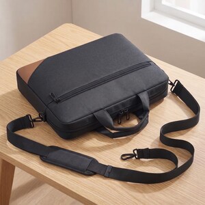Puede incluir: Bolso negro para portátil con un acento marrón en la esquina superior izquierda. El bolso tiene cierre de cremallera, asa y bandolera desmontable. El bolso está sobre una mesa de madera.