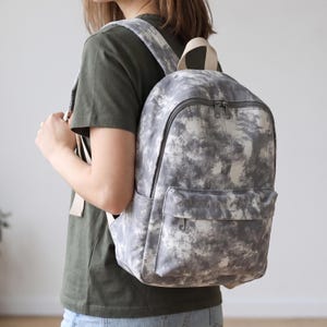 Puede incluir: Una mochila con un patrón de teñido anudado en gris y blanco, que cuenta con un compartimento principal grande, un bolsillo frontal más pequeño con cremallera y correas de hombro de color beige claro, cremalleras gris oscuro y asa superior.