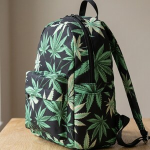 Puede incluir: Mochila negra con un estampado repetido de hojas verdes de cannabis. La mochila tiene un bolsillo frontal, un asa superior y correas ajustables. La mochila está sobre una superficie de madera.