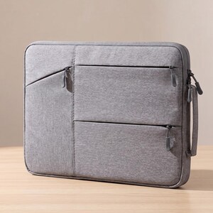 May include: A grey laptop sleeve with multiple zipped compartments. The sleeve has a textured fabric and a handle for carrying. The design includes a front pocket and two additional zipped sections. The overall aesthetic is minimalist and functional.
