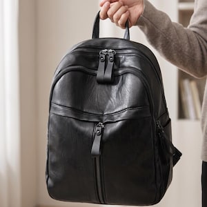 Puede incluir: Una mochila negra hecha de un material liso similar al cuero. La bolsa cuenta con un asa superior, dos compartimentos principales con cremallera y tiradores dobles, y un bolsillo vertical con cremallera en la parte frontal. Incluye bolsillos laterales.