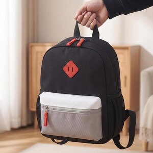 Puede incluir: Mochila negra con parche rojo en forma de diamante y tiradores de cremallera rojos. Cuenta con un bolsillo delantero blanco y gris y un bolsillo lateral de malla. La mochila está sujeta por una mano.