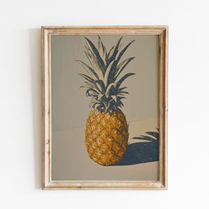 Op de afbeelding: Een ingelijste vintage print van een ananas. De ananas is weergegeven in tinten goud en bruin, met een gedetailleerde textuur. De bladeren zijn donkerblauw-grijs. De print zit in een lichtgekleurde, verweerde houten lijst.