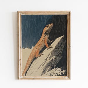 Puede incluir: Impresión vintage enmarcada de un lagarto. La obra presenta un lagarto naranja posado sobre una roca, sobre un fondo azul oscuro y negro. El lagarto está detallado con escamas texturizadas. El marco es de madera clara y envejecida.