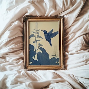 Peut inclure: Une impression encadr&eacute;e repr&eacute;sentant un colibri bleu en vol, approchant une plante aux grandes feuilles et aux fleurs d&eacute;licates. L'&oelig;uvre est pos&eacute;e sur un fond cr&egrave;me et encadr&eacute;e en or. L'esth&eacute;tique g&eacute;n&eacute;rale est botanique et &eacute;l&eacute;gante.