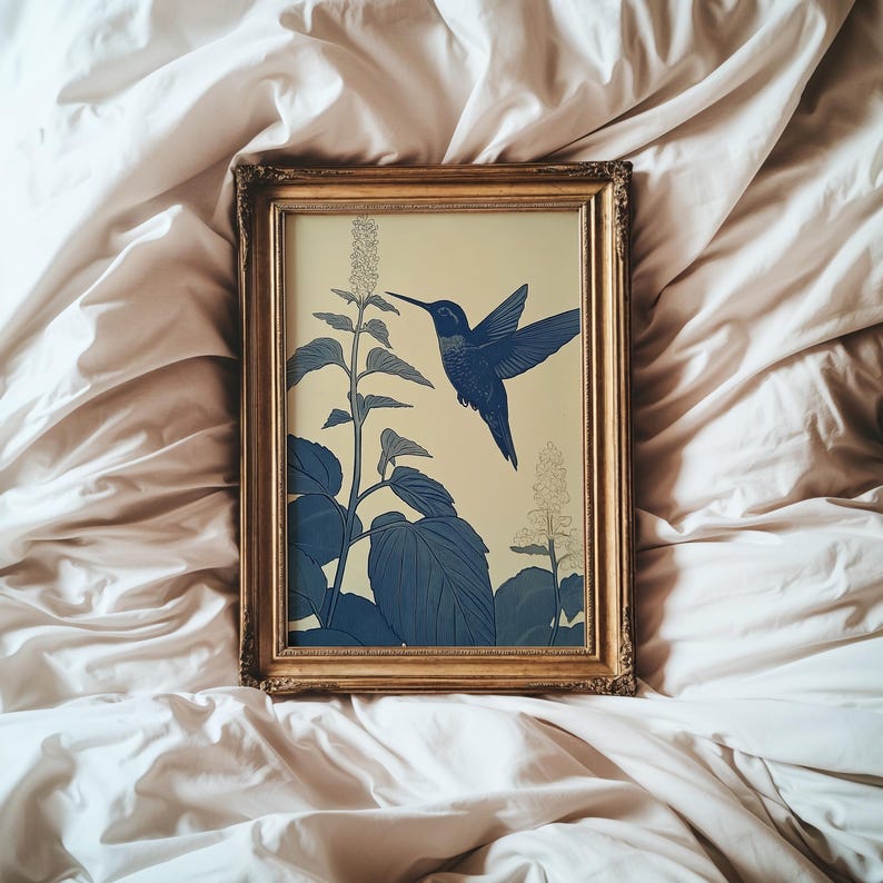 Peut inclure: Une impression encadr&eacute;e repr&eacute;sentant un colibri bleu en vol, approchant une plante aux grandes feuilles et aux fleurs d&eacute;licates. L'&oelig;uvre est pos&eacute;e sur un fond cr&egrave;me et encadr&eacute;e en or. L'esth&eacute;tique g&eacute;n&eacute;rale est botanique et &eacute;l&eacute;gante.