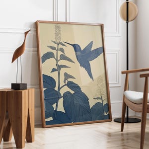 Peut inclure: Une affiche encadr&eacute;e repr&eacute;sentant un colibri bleu en vol, avec une illustration botanique de feuilles et de fleurs. L'&oelig;uvre d'art a un fond beige clair et est encadr&eacute;e dans un cadre en bois. L'affiche est appuy&eacute;e contre un mur blanc.