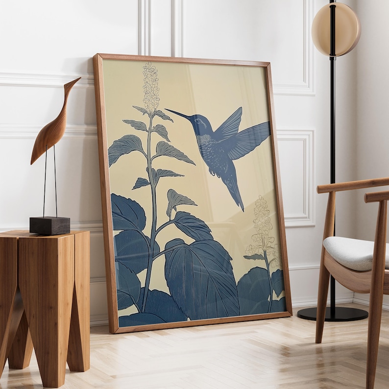 Peut inclure: Une affiche encadr&eacute;e repr&eacute;sentant un colibri bleu en vol, avec une illustration botanique de feuilles et de fleurs. L'&oelig;uvre d'art a un fond beige clair et est encadr&eacute;e dans un cadre en bois. L'affiche est appuy&eacute;e contre un mur blanc.