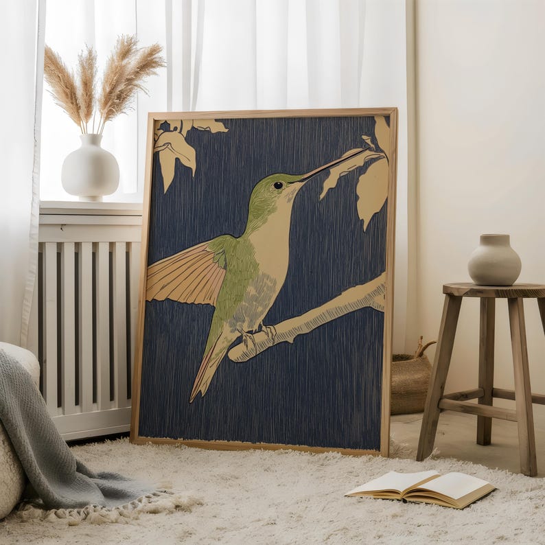 May include: A framed art print featuring a green and beige hummingbird perched on a branch against a dark blue background. The artwork is in a wooden frame and is displayed in a room with a white rug and a vase of dried flowers.