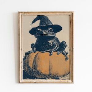 May include: Framed print of a black frog in a witch's hat, sitting on an orange pumpkin. The artwork has a vintage style, with a neutral background and a wooden frame. A whimsical Halloween-themed illustration.