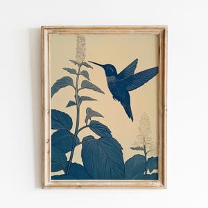Peut inclure: Une impression encadr&eacute;e repr&eacute;sentant un colibri bleu en vol, approchant une plante &agrave; fleurs. L'&oelig;uvre d'art a une esth&eacute;tique vintage, avec un fond cr&egrave;me et un cadre en bois brun clair vieilli. Le colibri et la plante sont rendus en nuances de bleu.