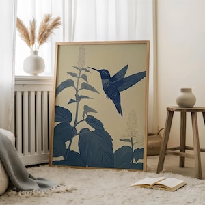 Peut inclure: Une affiche encadr&eacute;e repr&eacute;sentant un colibri bleu en vol, approchant une plante fleurie. L'&oelig;uvre d'art est pos&eacute;e sur un fond cr&egrave;me, avec la plante et l'oiseau rendus en nuances de bleu. Le cadre est en bois clair.