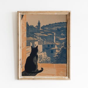 May include: A framed art print depicting a black cat observing a European village. The artwork uses blue, tan, and brown tones, showing detailed buildings and a distant tower. The cat is silhouetted against the view, creating a tranquil scene.