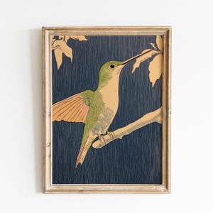 May include: A framed print featuring a hummingbird perched on a branch. The bird has green and tan plumage with orange wings, set against a dark blue background. The frame is a light, natural wood color.