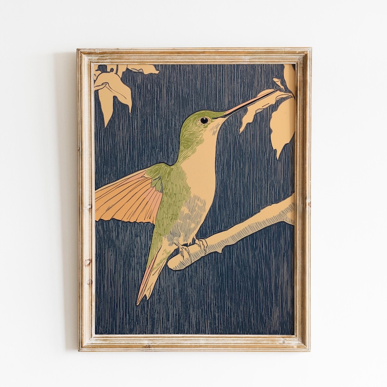 May include: A framed print featuring a hummingbird perched on a branch. The bird has green and tan plumage with orange wings, set against a dark blue background. The frame is a light, natural wood color.