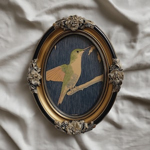 May include: Oval framed art print featuring a hummingbird perched on a branch. The bird has green and tan feathers with orange wings, set against a dark blue background. The frame is gold with black accents and floral embellishments.