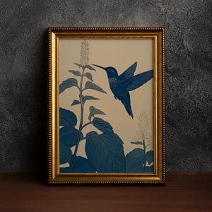 Peut inclure: Une impression d'art encadr&eacute;e repr&eacute;sentant un colibri bleu en vol, approchant une plante aux grandes feuilles et aux fleurs d&eacute;licates. L'&oelig;uvre d'art est pos&eacute;e sur un fond cr&egrave;me, avec l'oiseau et la plante rendus dans des tons bleus. Le cadre est dor&eacute;.