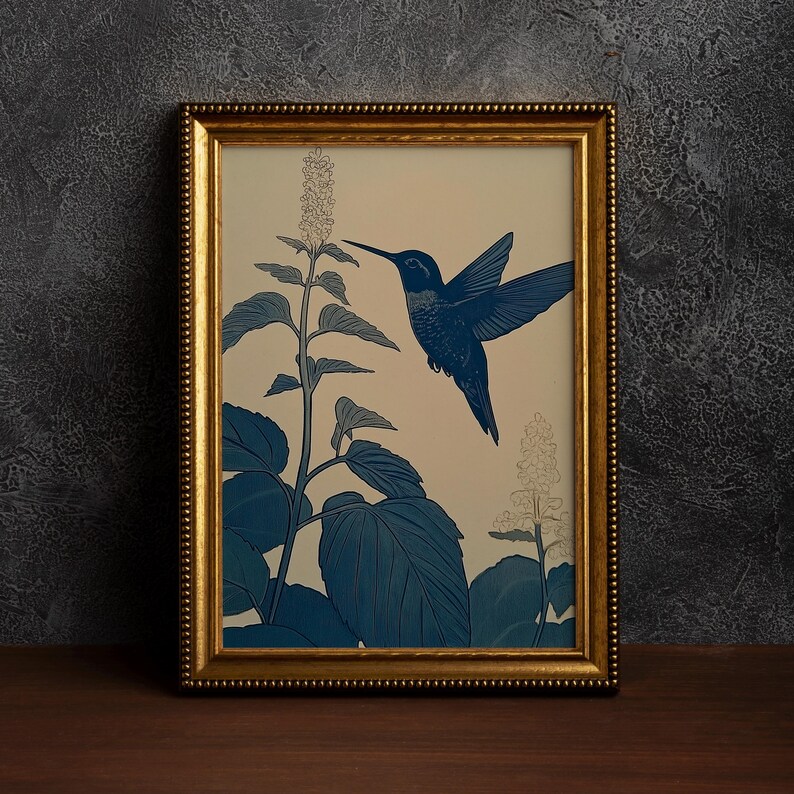 Peut inclure: Une impression d'art encadr&eacute;e repr&eacute;sentant un colibri bleu en vol, approchant une plante aux grandes feuilles et aux fleurs d&eacute;licates. L'&oelig;uvre d'art est pos&eacute;e sur un fond cr&egrave;me, avec l'oiseau et la plante rendus dans des tons bleus. Le cadre est dor&eacute;.