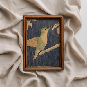 May include: A framed print featuring a hummingbird perched on a branch. The bird has green and tan plumage, with wings slightly spread. The background is a dark blue, and the frame is made of wood.