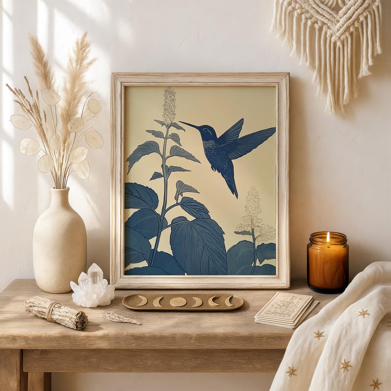 Peut inclure: Une impression encadr&eacute;e repr&eacute;sentant un colibri bleu en vol, sur fond cr&egrave;me avec des illustrations botaniques bleues. L'image est dans un cadre en bois clair, sur une table en bois avec un vase, des cristaux et une bougie.