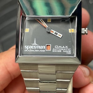 Op de afbeelding: Een zilverkleurig Spaceman OMAX automatisch horloge met een vierkante wijzerplaat en een zwarte wijzerplaat. Het horloge heeft een datumweergave en een zilveren band. De wijzerplaat heeft oranje en witte wijzers en markeringen. Het horloge is Swiss Made. De band is ongeveer 20 cm lang.