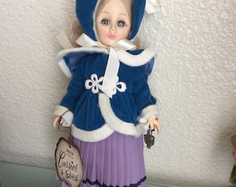 May include: A porcelain doll wearing a blue coat with white fur trim, a white ribbon, and a purple skirt. The doll is holding a pair of silver skates and has a tag that reads "Shatter Proof, Currier & Ives".