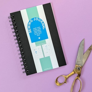 May include: A spiral-bound sewing journal with a black and mint green cover featuring the text Sewing Journal, track your makes and remember what (not) to do next time, and a pair of gold-toned metal sewing scissors with silver blades.