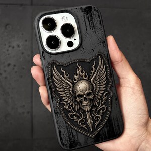 May include: A black smartphone case featuring a central embossed emblem of a winged skull atop a dagger, set within a shield-shaped border. The background of the case has a distressed, weathered texture with dark, scratched, and worn-in details.