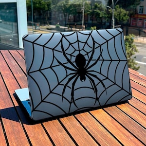 May include: A laptop computer with a gray case featuring a black spider and web design. The spider is centered on the case, with the web radiating outwards. The laptop is open and resting on a wooden table.